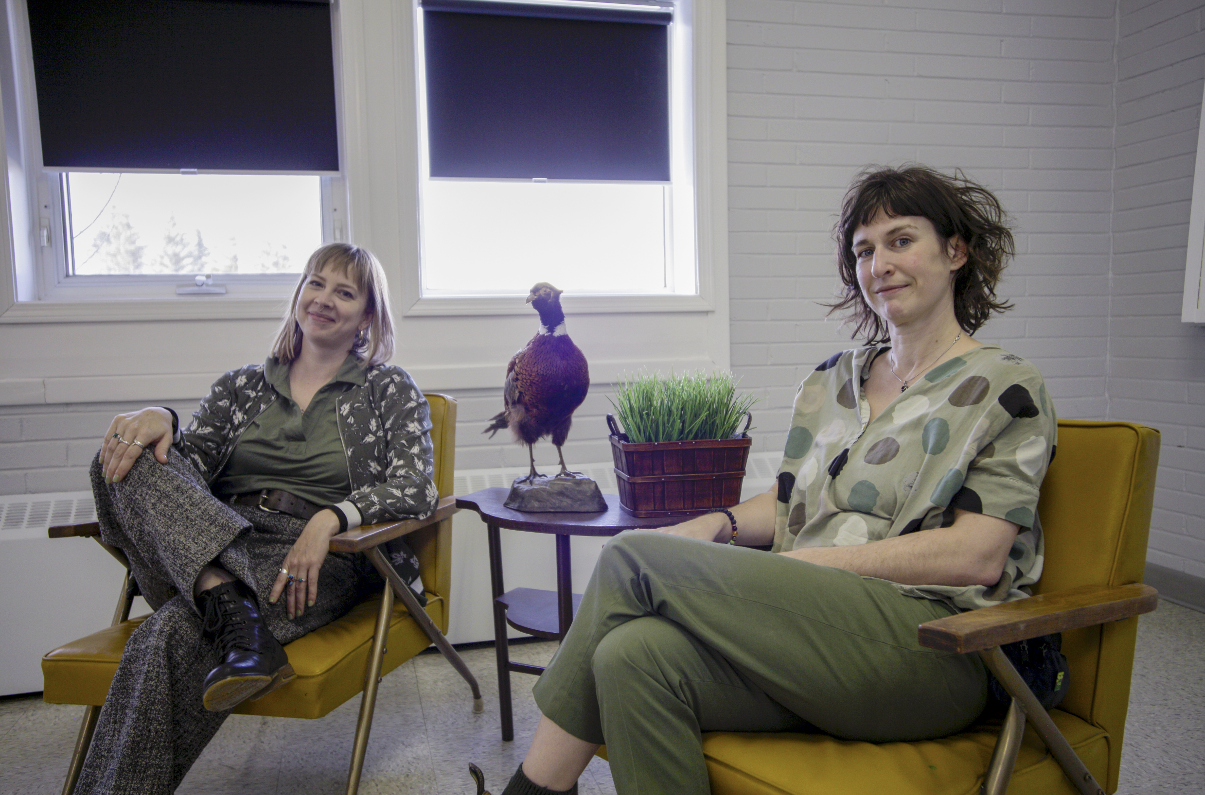 Two women sit in yellow armchairs in a bright room with white brick walls and windows behind them. A small table between them holds a decorative basket of grass and a mounted pheasant. The woman on the left wears patterned trousers, a green shirt, and a floral jacket. The woman on the right wears green pants and a polka-dotted shirt. Both are relaxed and smiling slightly, creating a casual and welcoming atmosphere.